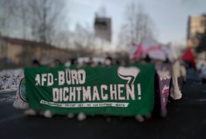 Fronttransparent der Demonstration mit der Aufschrift "AfD-Büro dichtmachen, das freundliche Gesicht des NS aus Dortmund vertreiben." Die dahinter stehenden Demonstrant:innen sind zur unkenntlichmachung verschwommen.