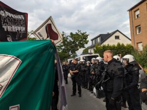 Demonstrant:innen hinter einem Grünen Transparent, über ihnen Fahne und Doppelhalter mit der aufschrift "Offenes Antifa Treffen Dortmund", daneben eine Reihe Bereitschaftspolizist:innen