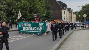 Eine Demonstration, Blick von vorne auf ein grünes Transparent mit der Aufschrift "AfD-Büro dichtmachen - Das freundiche Gesicht der NS aus Dortmund werfen", dazu Fahnen und daneben eine Reihe Bereitschaftspolizist:innen