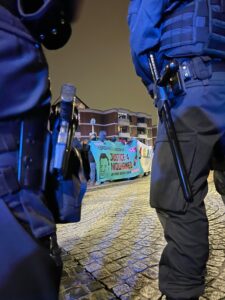 Die Zwischenkundgebung an der Wache Nord, Blick auf die Demo zwischen zwei Bereitschaftspolizist:innen hindurch