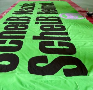 Bild von einem Transparent das auf dem Boden liegt, daneben eine Schale mit Pinseln. Auf dem Transparent steht "Scheiß Nazis. Scheiß Heimat.", dazu ein Antifalogo