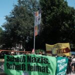 Anblick einer Demonstration von vorne. Auf dem grünen Fronttransparent ist die Aufschrift "Scheiß Nazis. Scheiß Heimat." sowie ein leicht angeschnittenes Antifalogo am unteren Rand zu sehen. Ein weiteres Transparent an der linken Seite der Demo trägt die Aufschrift "The future is stilll unwritten", ein Hochtransparent im Hintergrund den Text "never mind Germany, here is the Antifa". Die Gesichter der Demonstrant:innen hinter den Transparenten sind verpixelt.