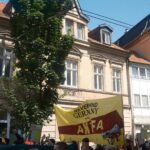 Anblick einer Demonstration von der Seite. Mittig ein Hochtransparent mit der Aufschrift "never mind Germany, here is the Antifa". Die Gesichter der Demonstrant:innensind verpixelt. Im Vordergrund sind 3 Polizist:innen am Rande der Demonstration zu sehen.